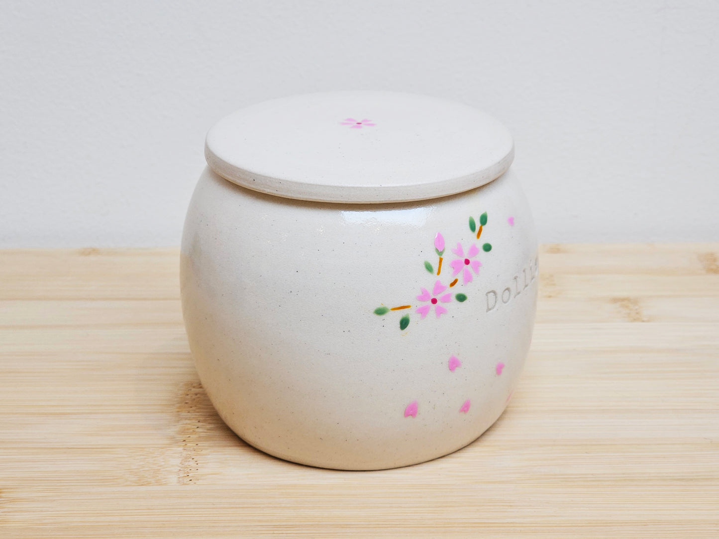 Right side view of a handmade ceramic pet urn featuring hand-painted cherry blossoms and drifting petals. Personalized with the engraved name "Dollie." This custom memorial urn is ideal for honoring a beloved pet, combining artisan craftsmanship with a nature-inspired cherry blossom design.