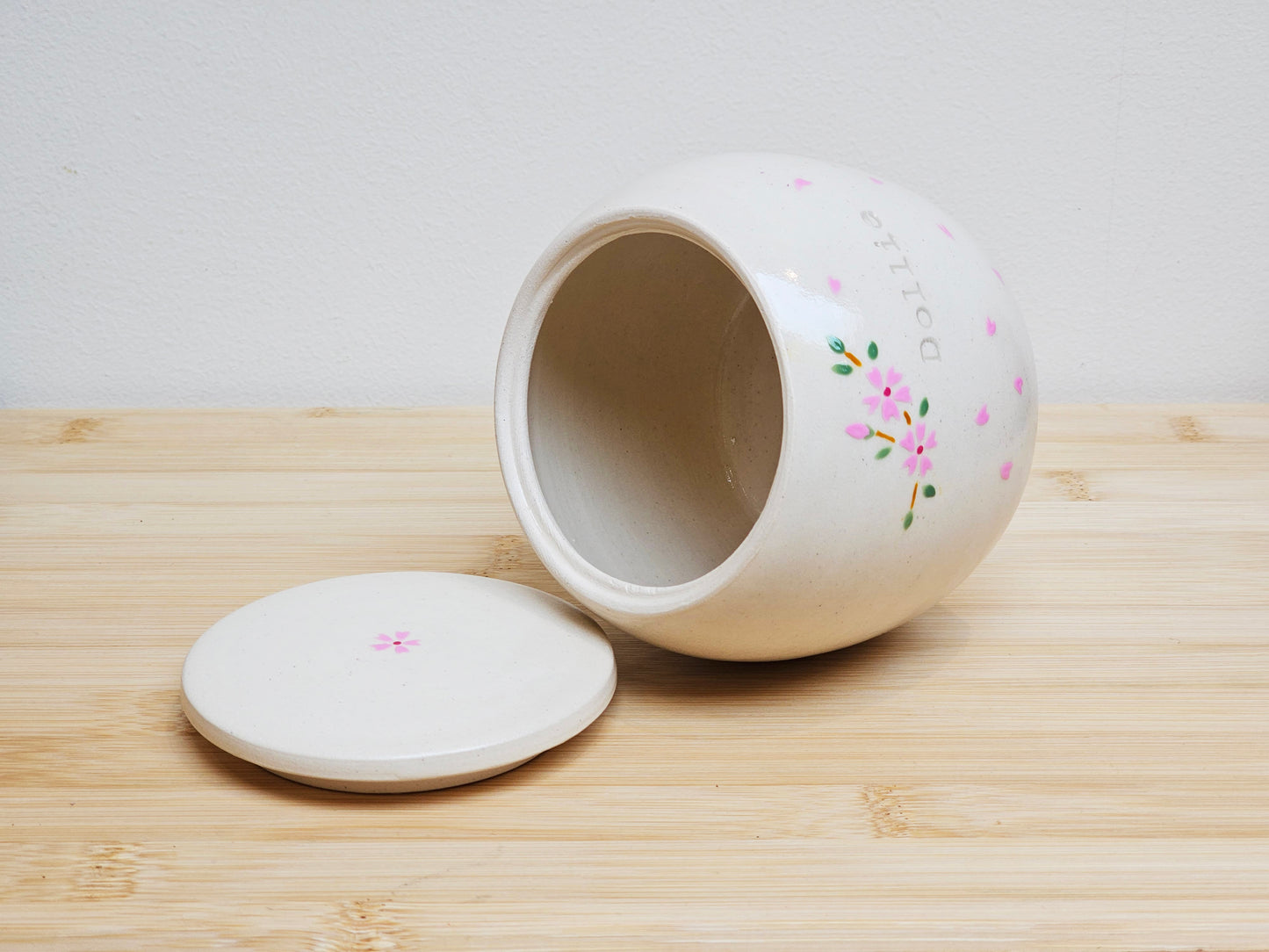 Opened view of a handmade ceramic pet urn with the lid removed, revealing the smooth interior. The urn features a delicate cherry blossom design with drifting petals and is personalized with the name "Dollie." A unique and elegant memorial urn for a beloved pet.