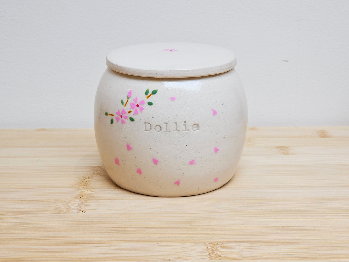 A handmade ceramic pet urn decorated with delicate cherry blossom branches and drifting petals. The soft pink blossoms stand out against the smooth, light-toned surface. The urn is engraved with the name "Dollie," adding a personal, heartfelt touch.