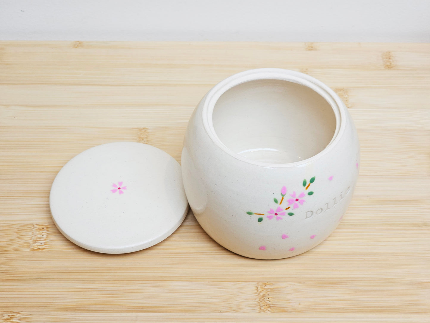 Top view of a handmade ceramic pet urn with the lid removed, showing the open interior. The urn is adorned with a cherry blossom design and drifting petals, and engraved with the name "Dollie." A beautifully crafted, personalised pet memorial urn.