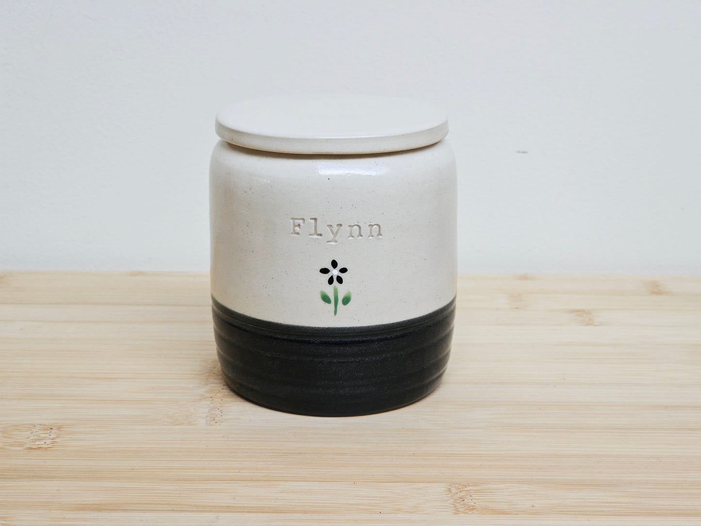 Custom pet urn with 'Flynn' and hand painted black flower, with a black base. Personalised pet memorial ceramic jar with a lid.