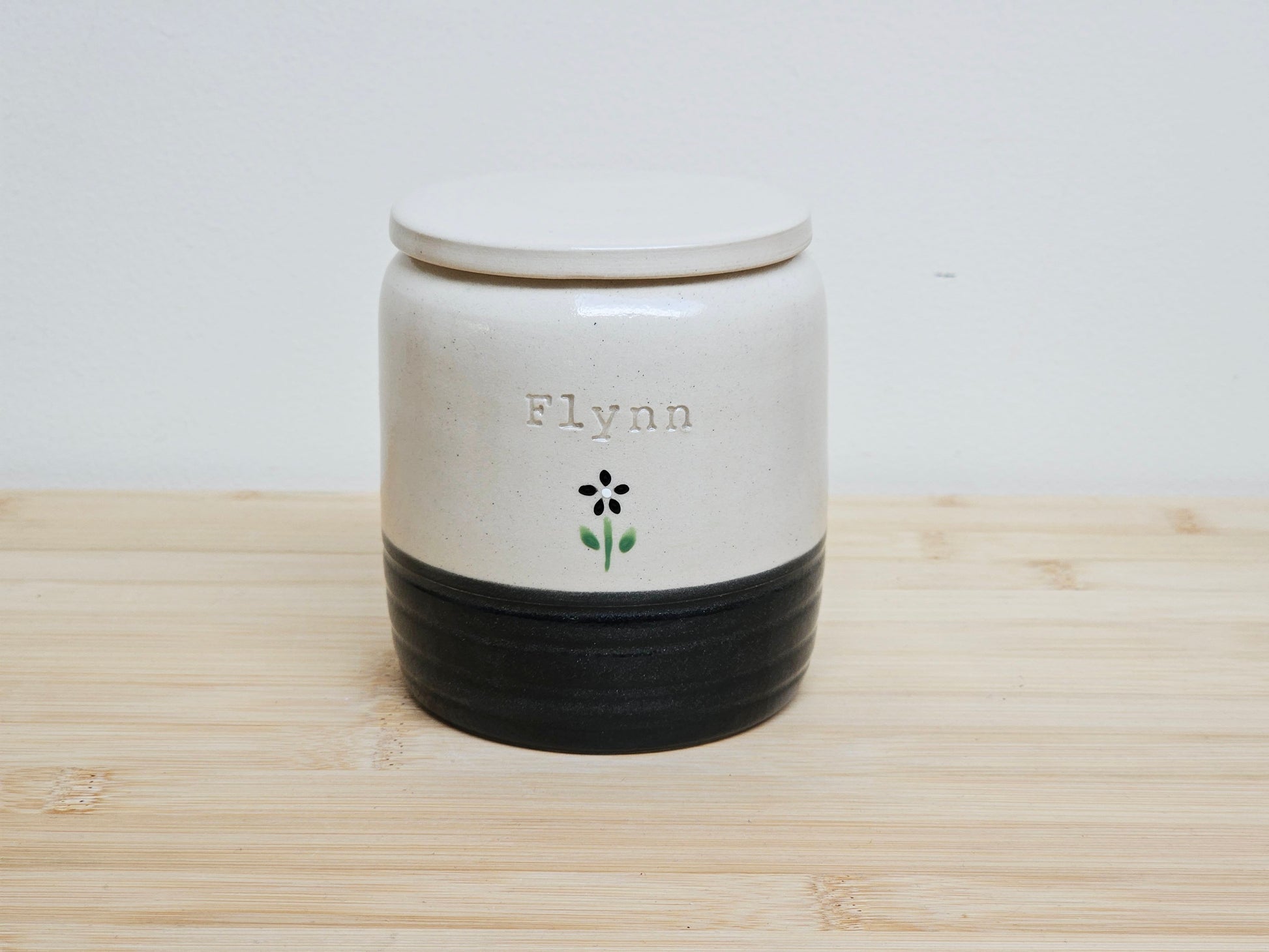 Custom pet urn with 'Flynn' and hand painted black flower, with a black base. Personalised pet memorial ceramic jar with a lid.