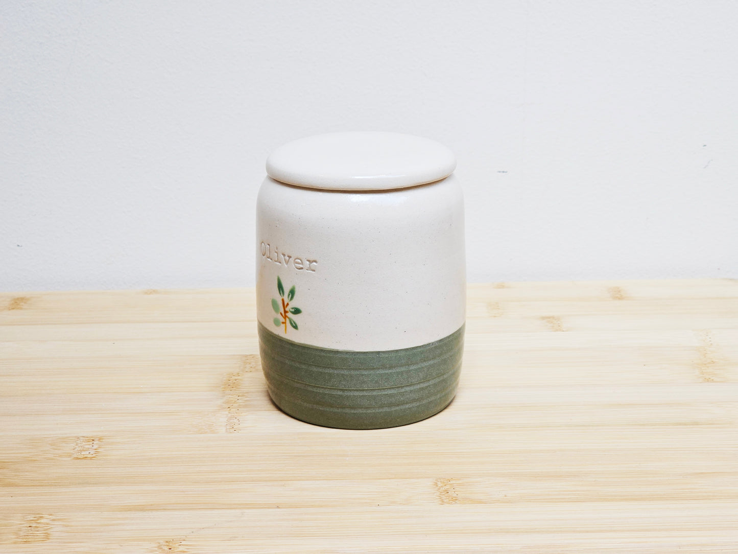 White ceramic urn with green base and 'Oliver' branding on a wooden surface.