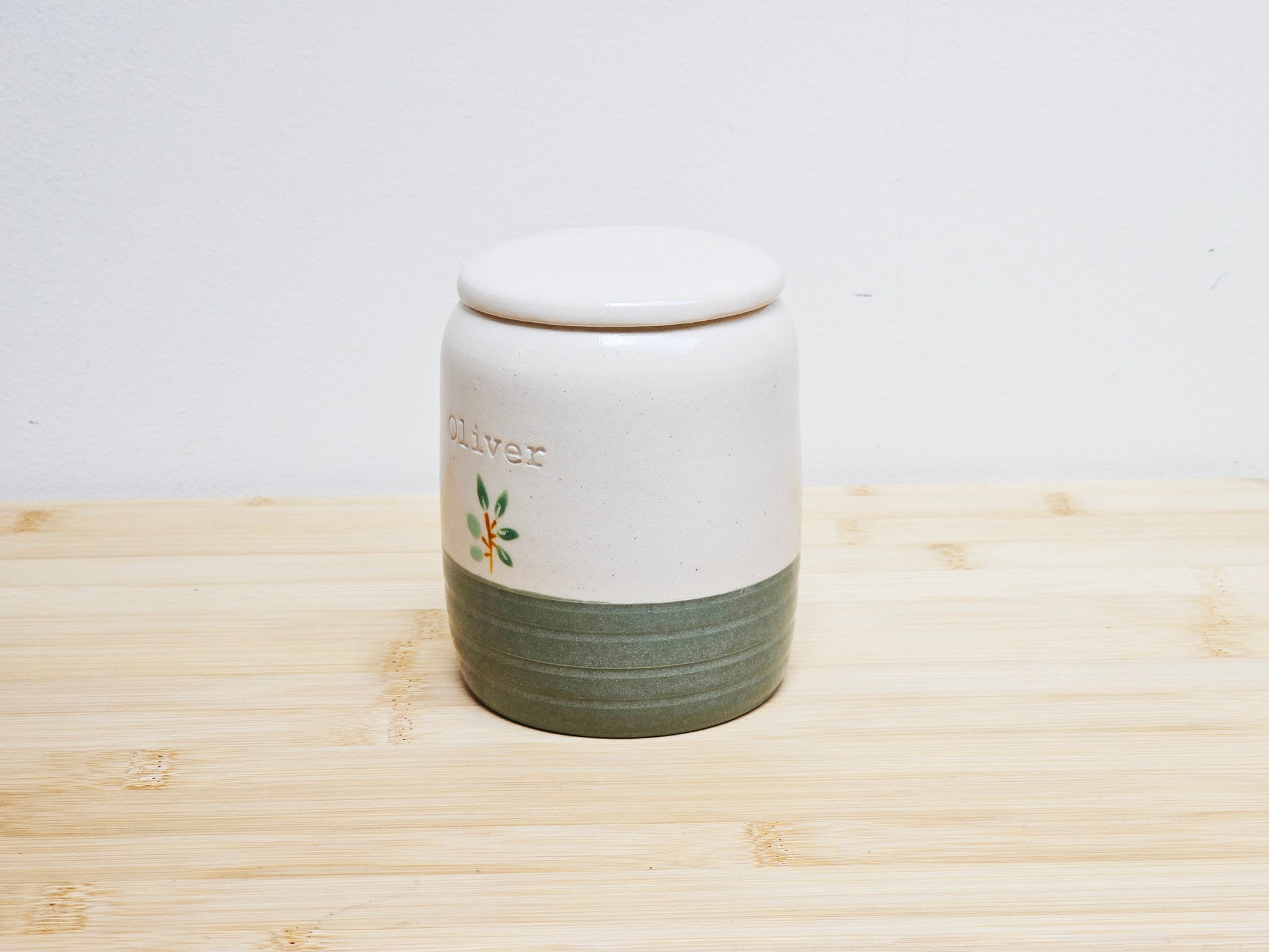 White ceramic urn with green base and 'Oliver' branding on a wooden surface.
