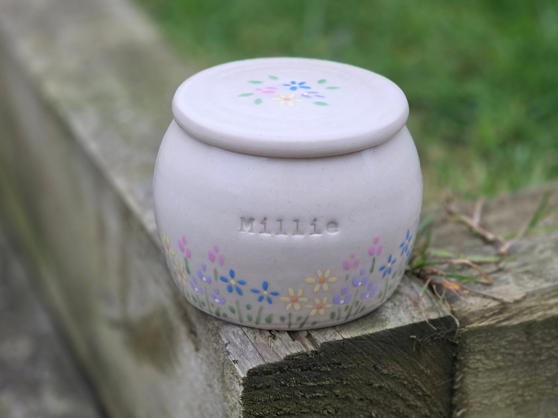 Small white ceramic jar with pastel floral designs and 'Millie' branding on a stone surface.